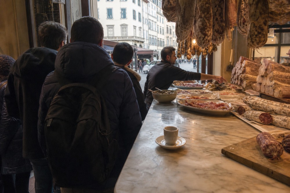 A small group standing at a Florence food counter while a guide points at trays of cured meats