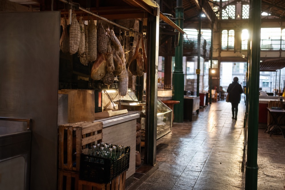 A covered market hall with food stalls and hanging cured meats under iron and glass