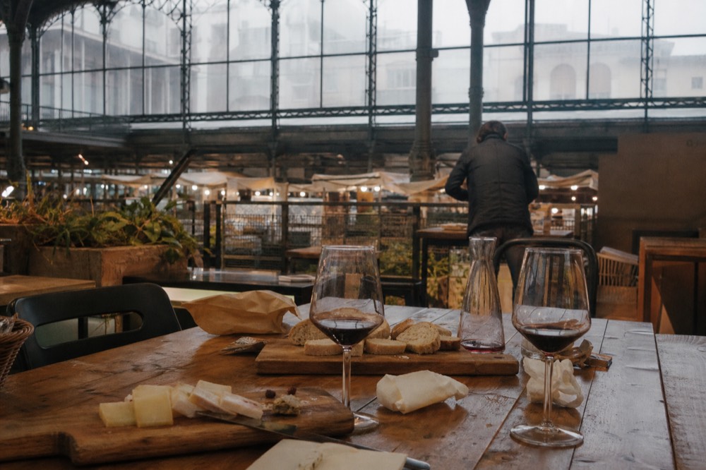 A long wooden table set with bread, cheese boards and glasses of red wine under warm interior light