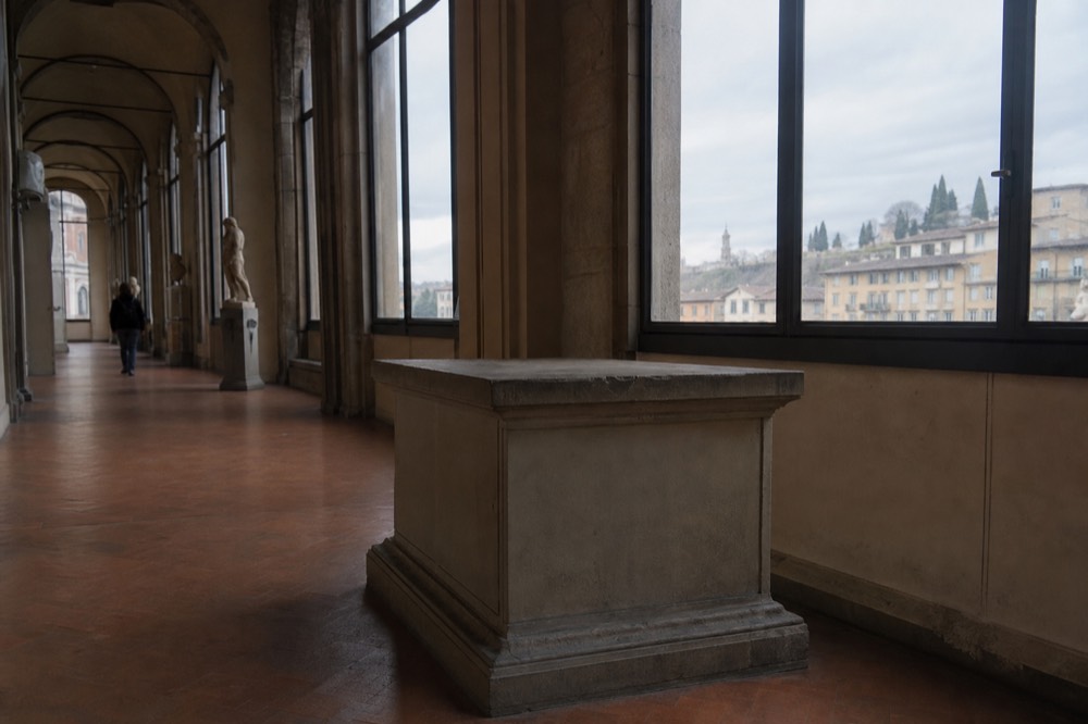 A quiet museum gallery corridor in Florence with tall arched windows and empty stone pedestals