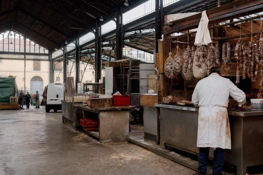 Sant'Ambrogio market interior with wooden stalls, hanging salami, and a butcher at work