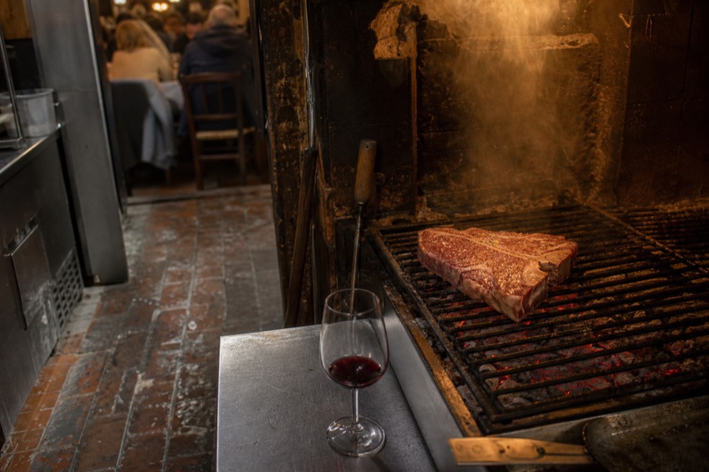 A thick T-bone steak resting on a grill over glowing coals in a Florentine kitchen