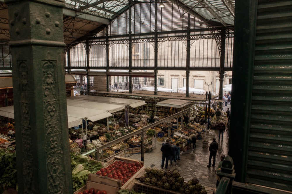 A covered historic market hall with produce stalls and shoppers under iron and glass