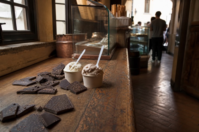 Artisan chocolate shards and two cups of gelato on a worn wooden counter inside a gelateria in Florence, Italy.