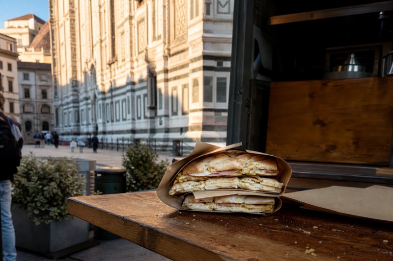Paper-wrapped schiacciata panini stacked on a wooden street-food counter in Florence, Italy, with the Duomo's marble facade blurred behind.