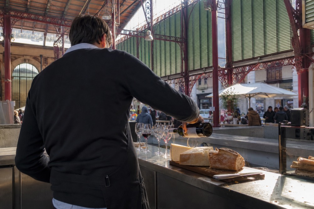 A sommelier pouring red wine into glasses on a market counter beside plates of cheese
