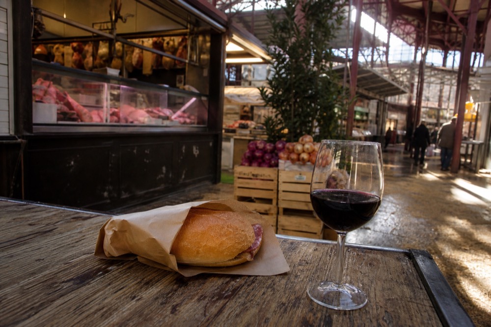 A paper-wrapped sandwich and a glass of red wine on a market stall counter