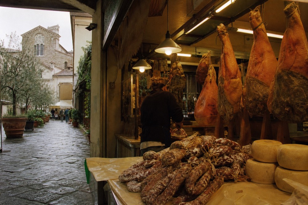 A market display of cured hams, salami and whole pecorino cheeses under lights