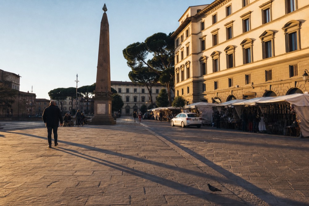 A sunlit Florentine piazza with a stone obelisk and market stalls in the distance