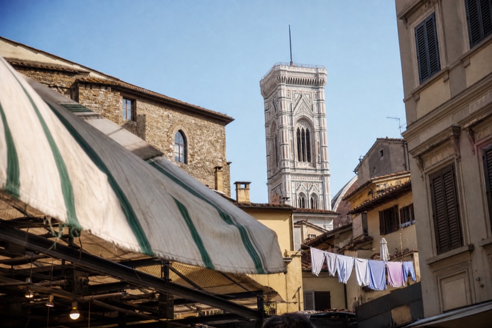 A Florence street with a market awning, a bell tower rising above the rooftops