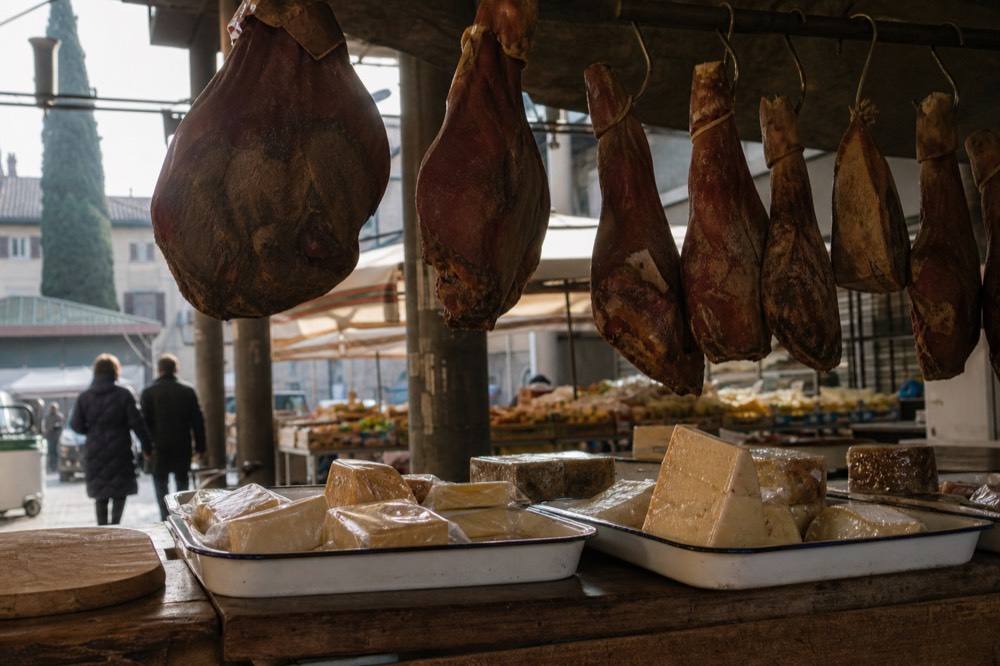 Close view of a Tuscan market counter with cured hams hanging above trays of cheese