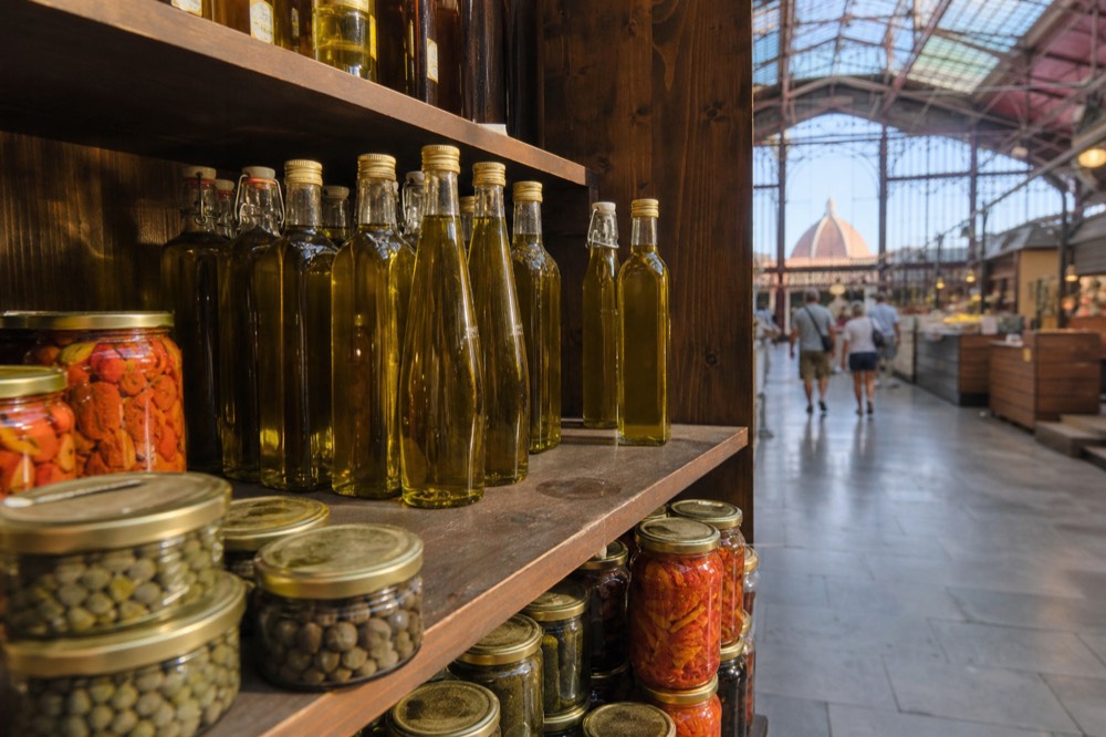 Bottles of golden olive oil and jars of preserves lined up on a wooden market shelf