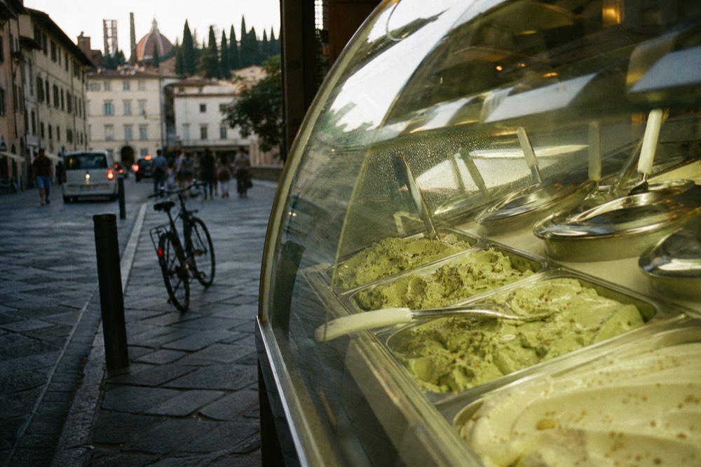A gelato counter with rows of tubs and a spatula resting on top
