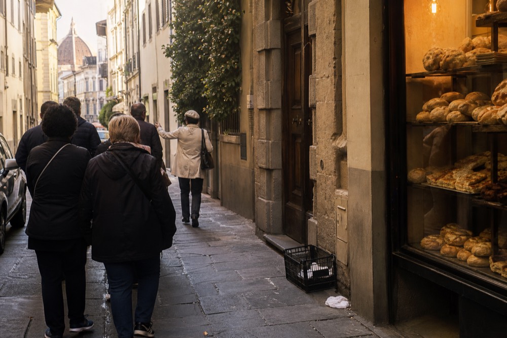 A narrow Florentine street with a guide leading a small group past a bakery display window filled with bread and pastries