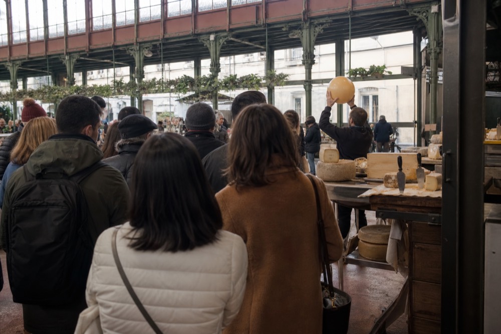 Inside the ground floor of Mercato Centrale with a food tour group gathered around a cheese and salumi stall, the guide holding up a wheel of pecorino