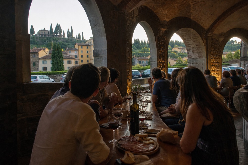 A food tour group seated at a long wooden table in a wine bar, glasses of red wine and plates of cheese and salumi in front of them