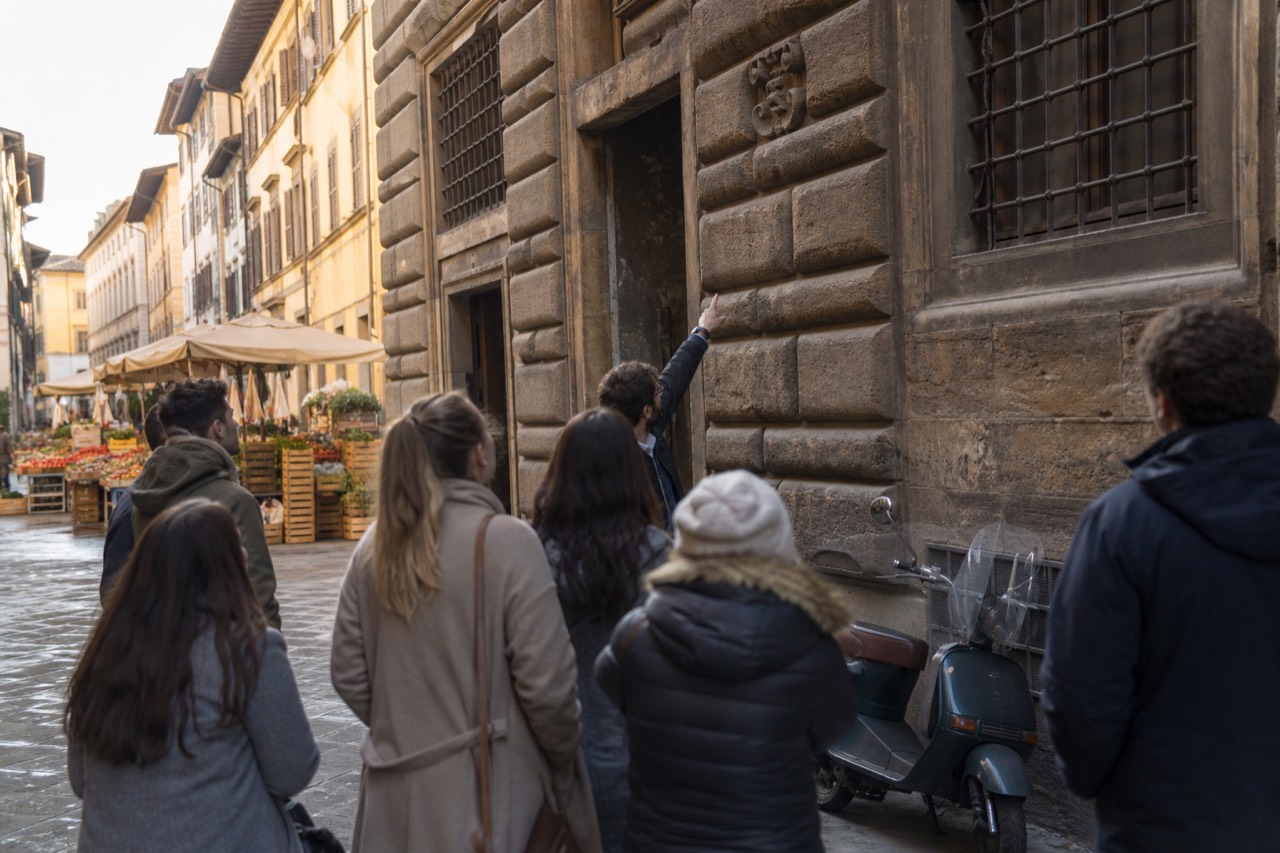 A walking food tour group in Florence passing a historic stone building with a guide pointing out a detail, market stalls visible in the background