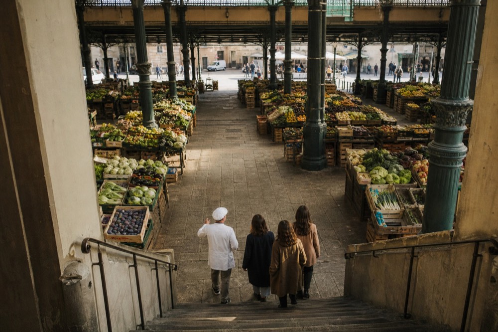 Chef guiding three visitors through a covered food market past produce stalls