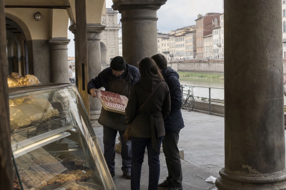 Local guide explaining a tray of cured meats to two visitors at a deli counter