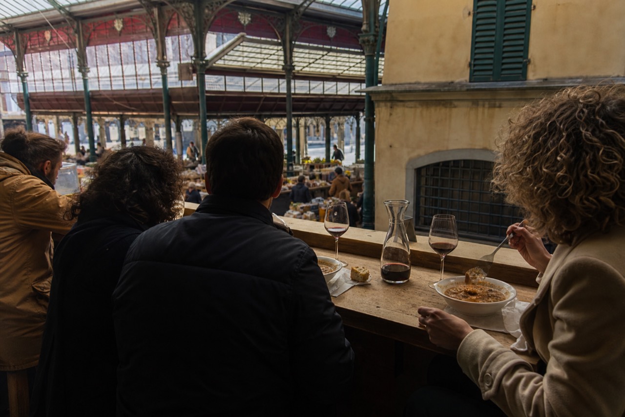 Small group of diners seated at a wooden counter with wine glasses and plates of Tuscan food